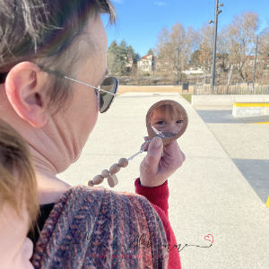 A white mum looks at her white toddler in a wooden portable rearview mirror, the child is carried in a handwoven wrap, they're outside.