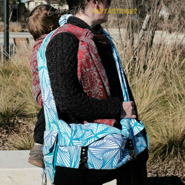 Blue babywearing bag in crossbody style, on the hip, baby on back. Picture taken outside.