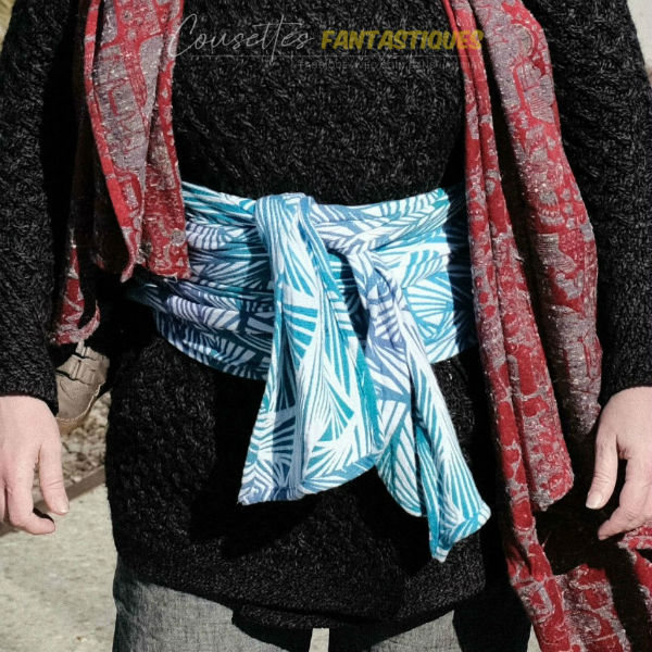 Sac de portage bleu en mode sac à dos, finition 'pagne/belly wrapping', bébé au dos. Photo prise en extérieur.