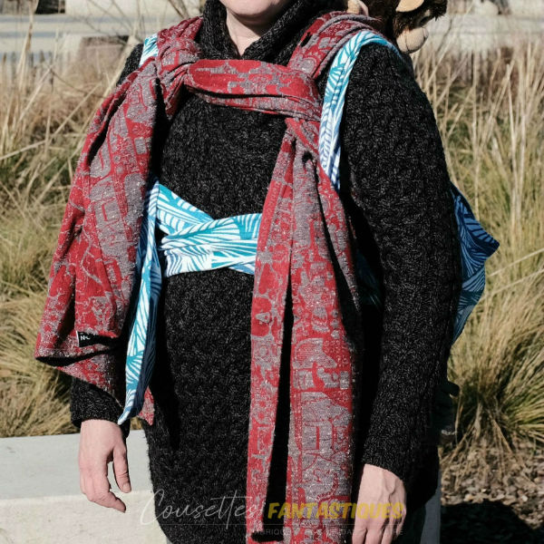 Blue babywearing bag in Backpack style, tibetan finish, baby on back. Picture taken outside.