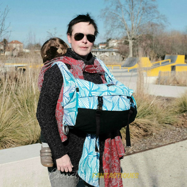 Blue babywearing bag in high front bag style, tied in front, baby on back. Picture taken outside.