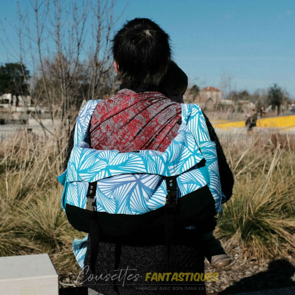Blue babywearing bag in backpack style, baby in front. Picture taken outside.