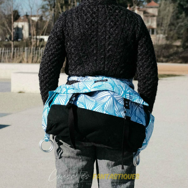 Blue babywearing bag in 'pagne' style without the rings, 'belly wrapping' finish, without baby, vue de dos. Picture taken outside.