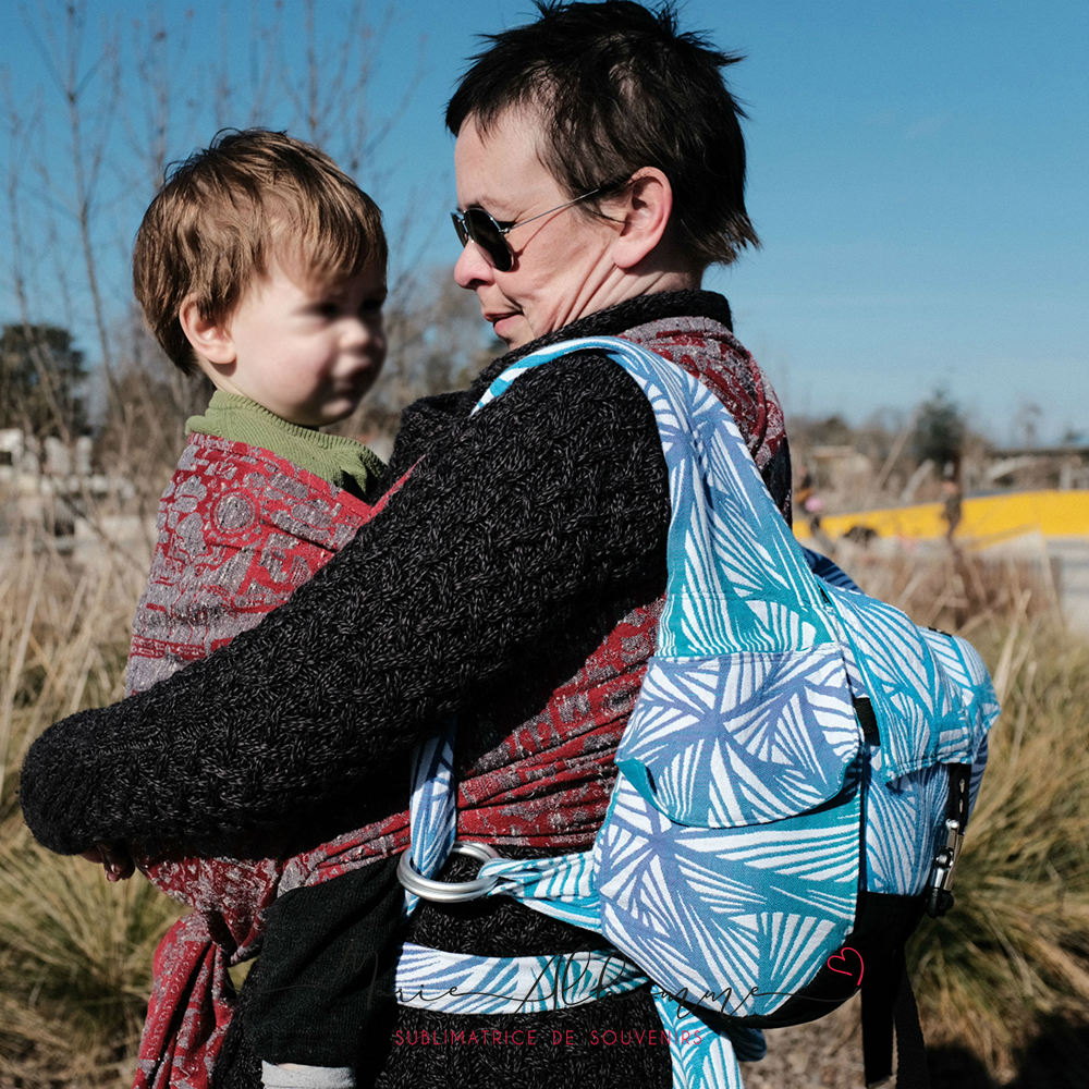 A white mum carries her white toddler in a red and grey wrap, and also carries a blue babywearing backpack, they're outside.